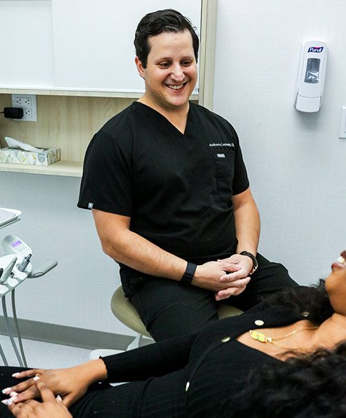 Smiling dentist in black scrubs chatting with a reclined patient before a night guard fitting in New York