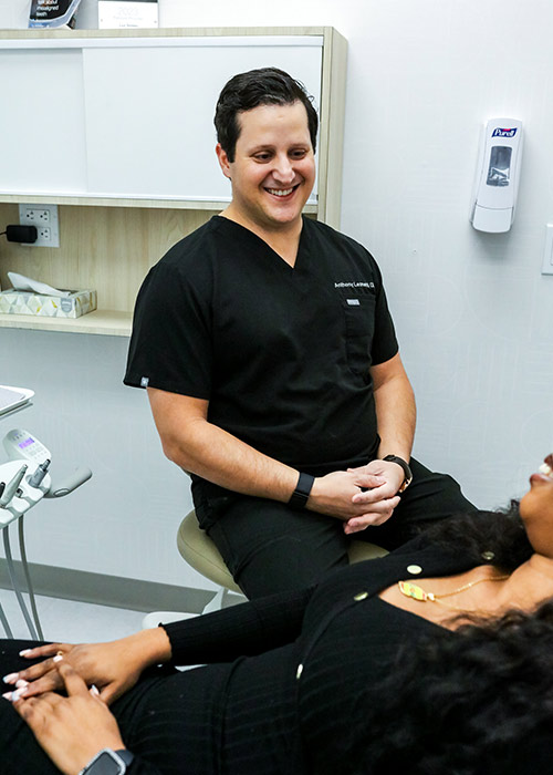 Smiling dentist in black scrubs chatting with a reclined patient before a night guard fitting in New York